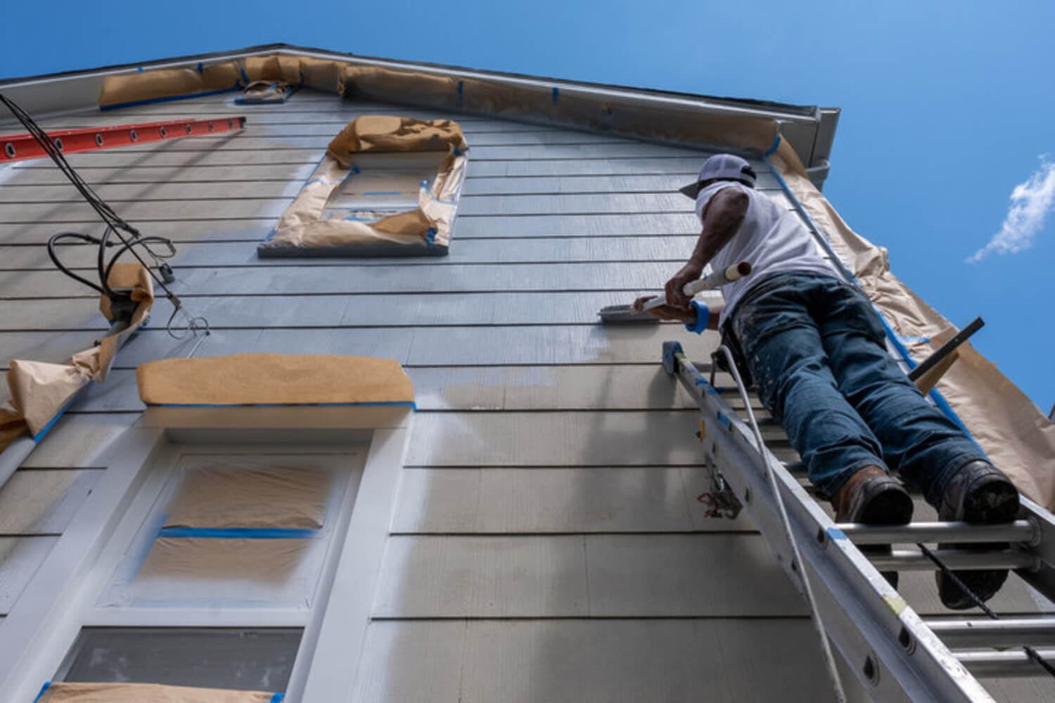 Painter on a ladder applying gray paint to the lap siding of a residential home on a clear summer day with windows masked for protection