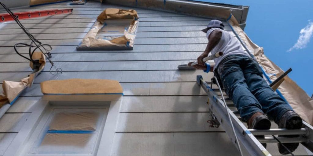 Painter on a ladder applying gray paint to the lap siding of a residential home on a clear summer day with windows masked for protection